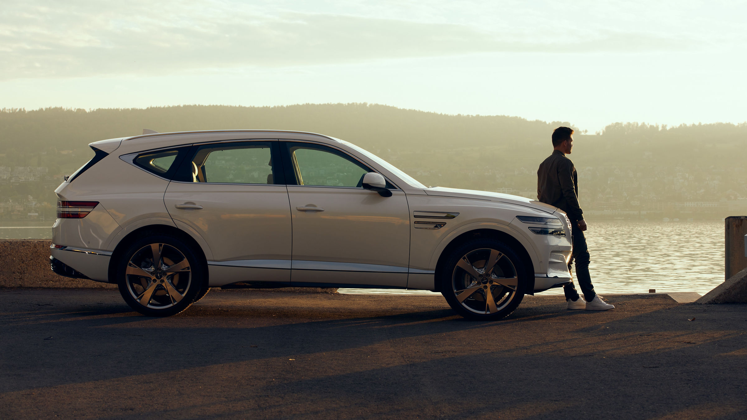 White Genesis GV80 at sunset by the sea with man leaning against bonnet