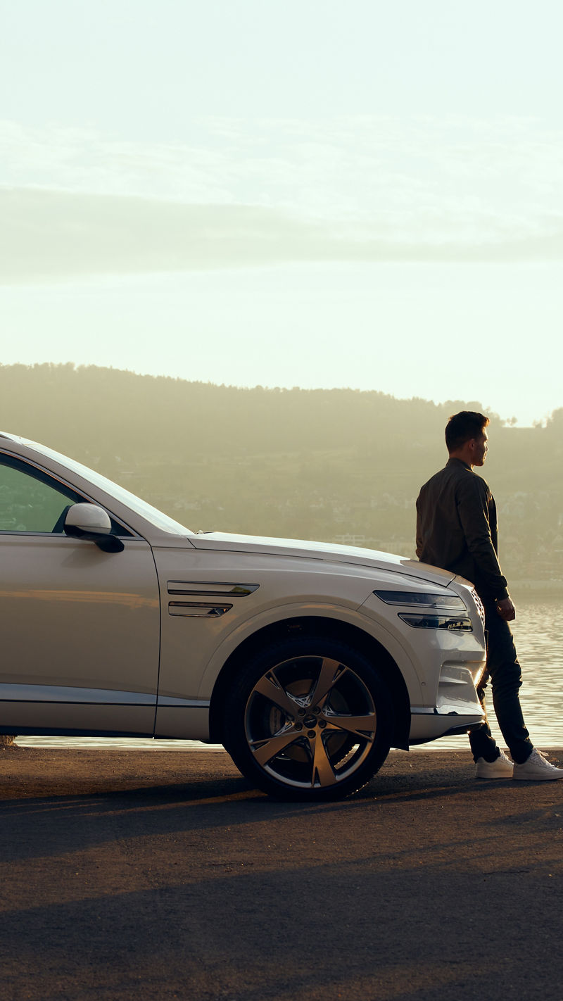 White Genesis GV80 at sunset by the sea with man leaning against bonnet