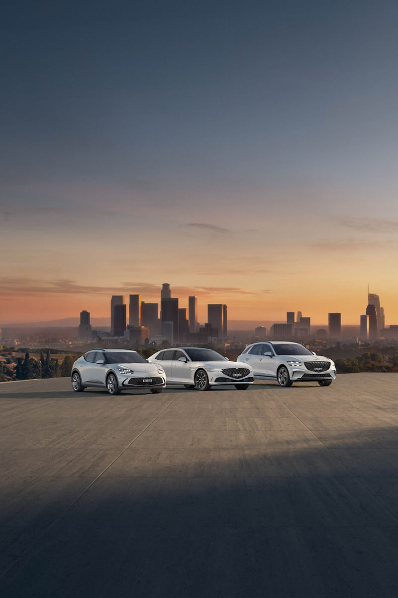 A white Genesis GV80 parked by the waterfront with a city skyline and bridge in the background.