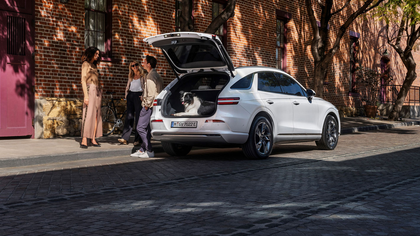 Three people stand next to a white Genesis Electrified GV70 with an open trunk and a dog in it