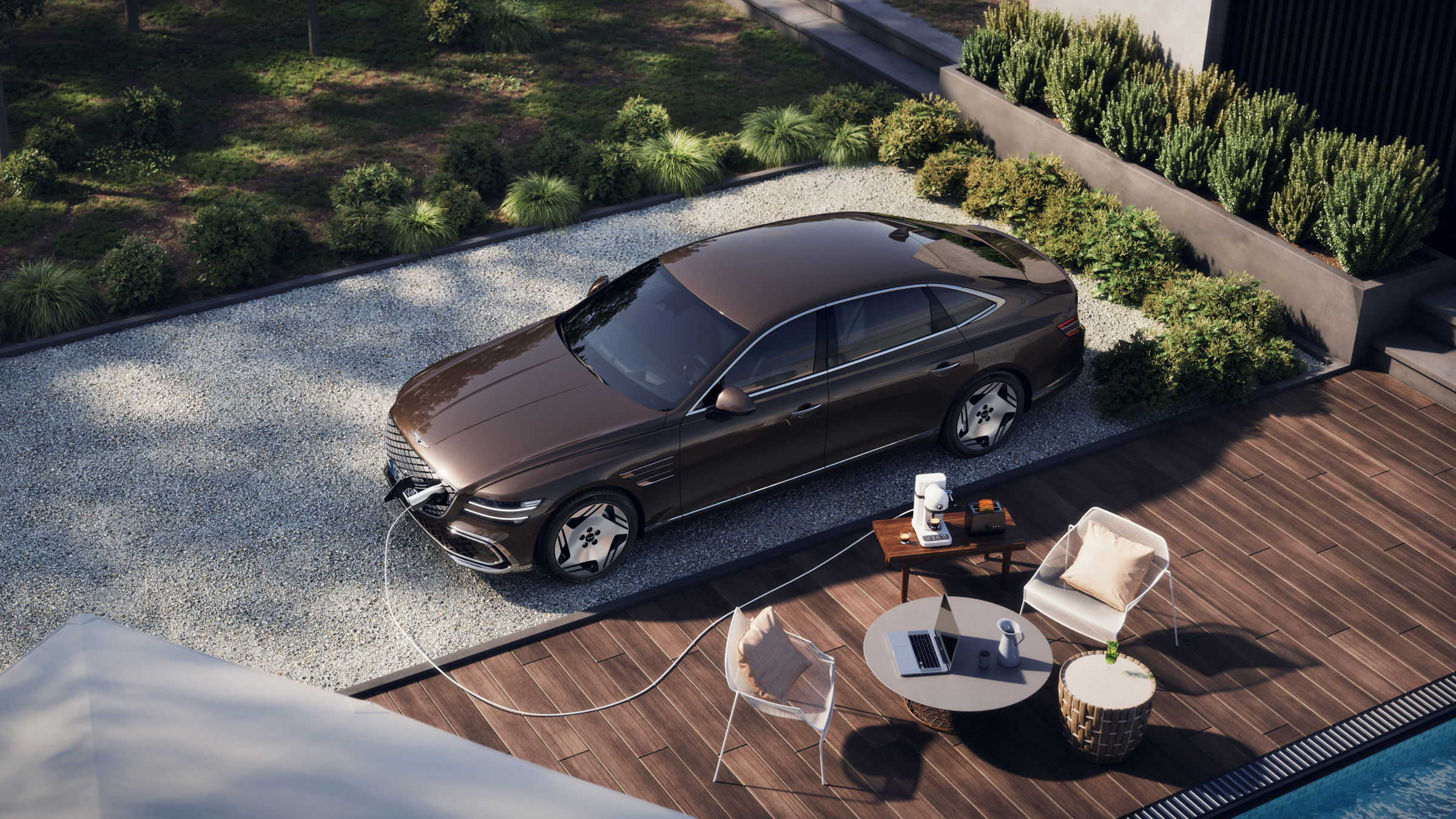 Electrified G80 wordt opgeladen in een tuin met grind. Voor de auto staan een laptop op een koffietafel, een kleine koffiemachine en meer, wat een ontspannen sfeer creëert.