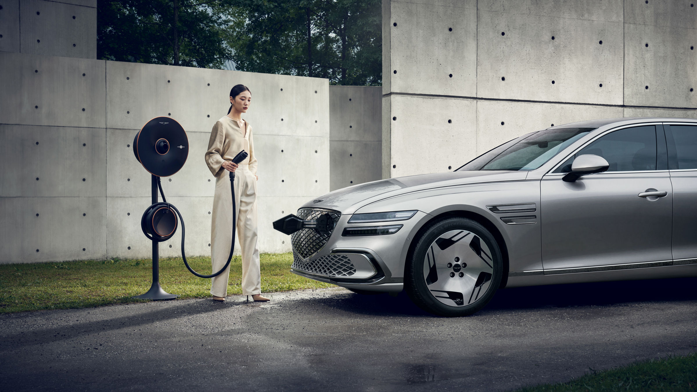An Electrified G80 is parked in an outdoor space, against an exposed concrete wall. A young woman is standing in front of the vehicle with a Genesis EV Home Charger in her hand.