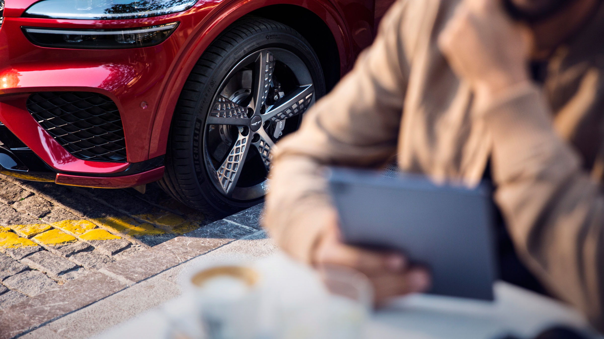 Tyres of a red Genesis GV70 Sport, in the foreground a person with a tablet and coffee