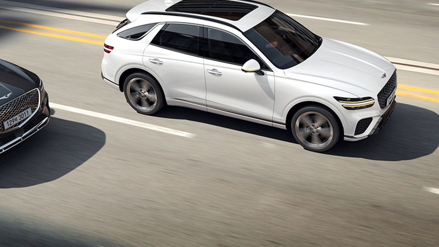 A white Genesis GV70 SUV driving on a highway, overtaking a gray car on the left lane.