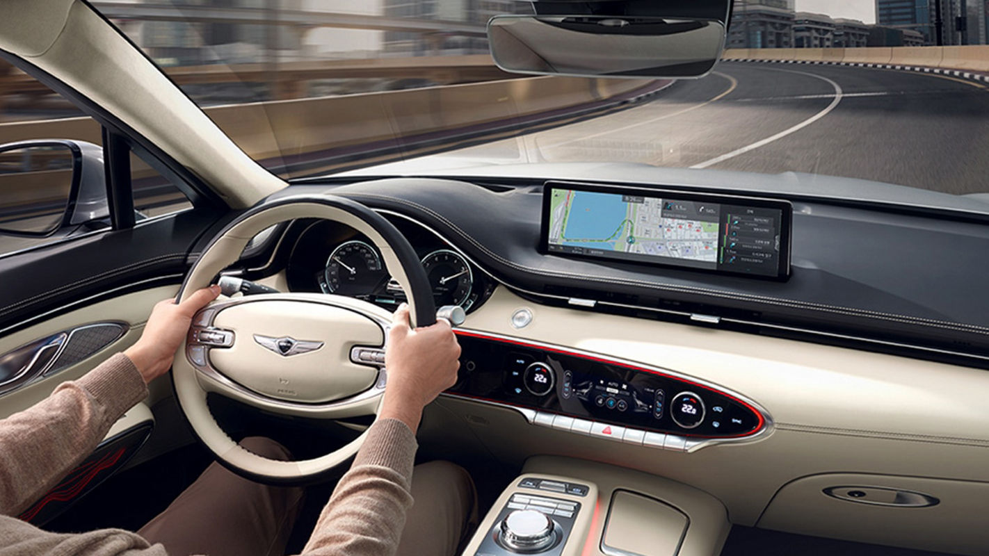 A luxury car interior of the Genesis GV70 featuring a white steering wheel, a touchscreen display showing navigation, and a curved dashboard design.