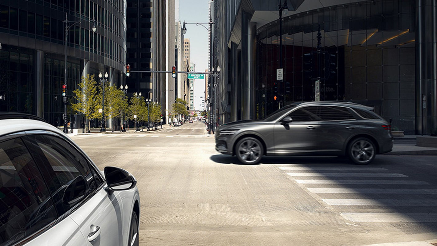 A silver Genesis GV70 SUV crossing a modern urban intersection with glass buildings in the background.