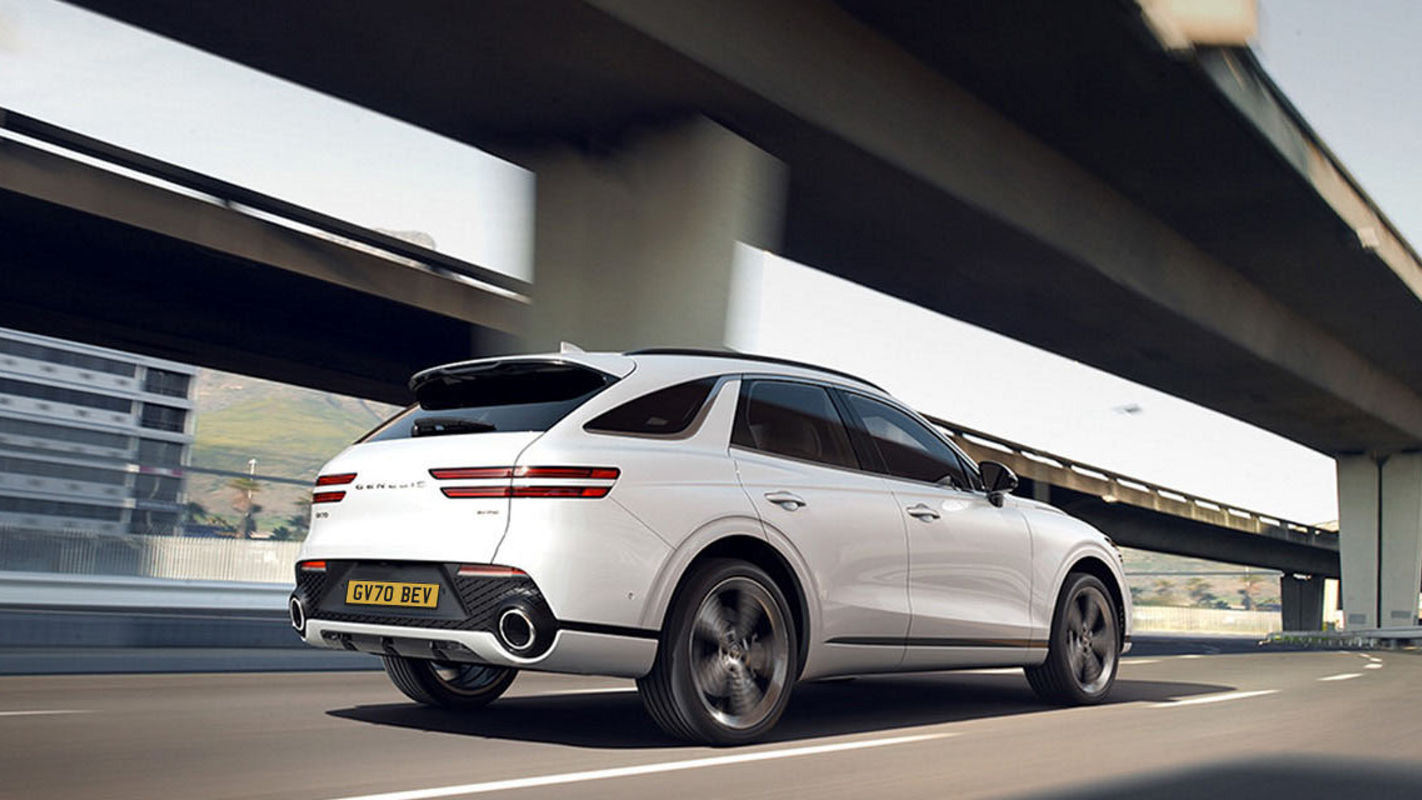 A rear view of a white Genesis GV70 SUV driving on a highway, showcasing its sleek tail design and dual exhausts.