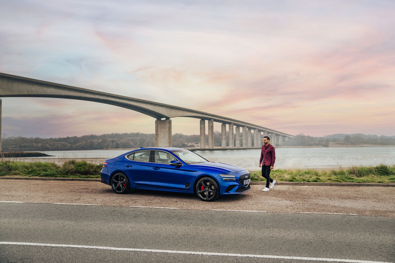 Genesis G70 blue exterior in front of a bridge - with man walking aside the car