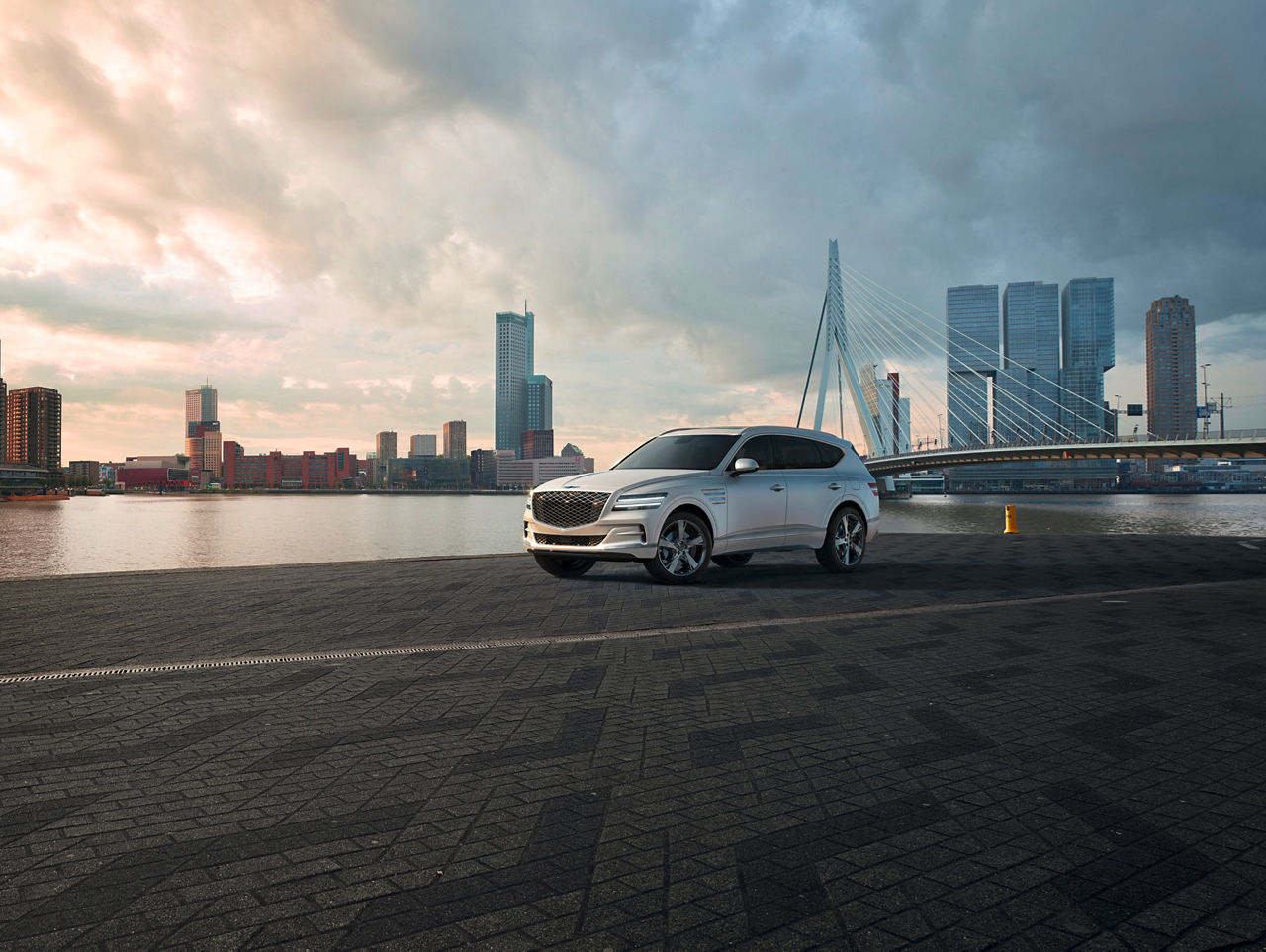 White Genesis GV80 in front of a river with a bridge and the skyline of a city in the background