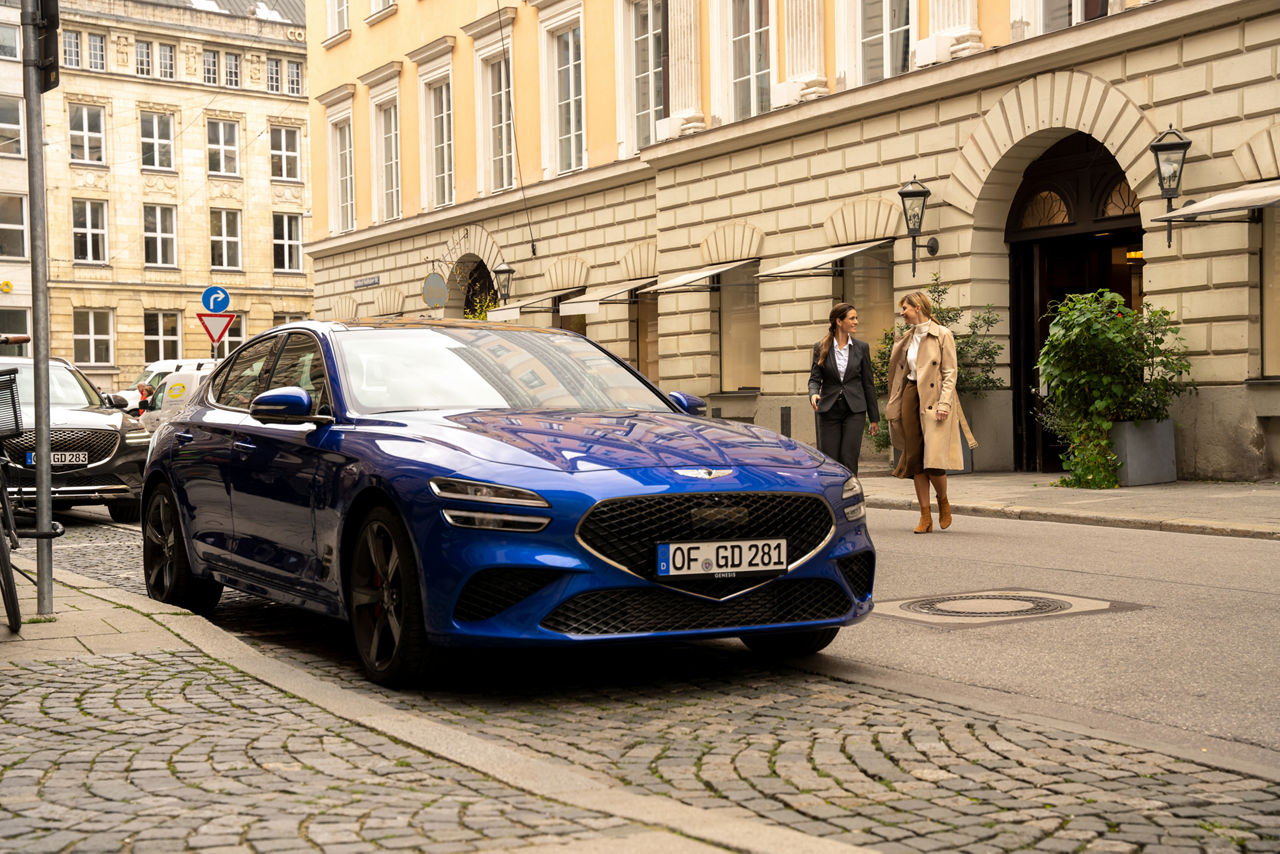Blue Genesis G70 parked at the edge of a road with buildings and people in the background