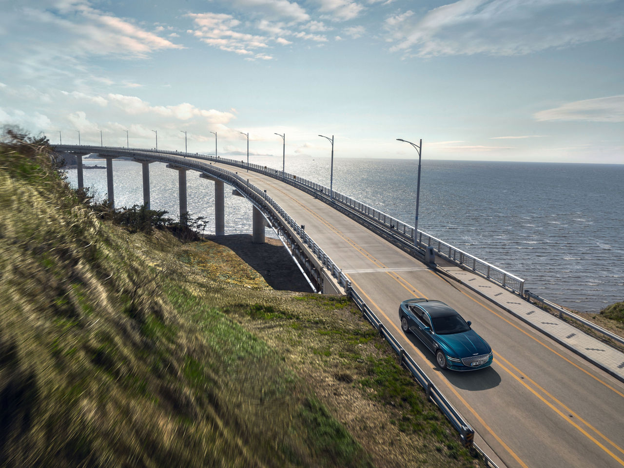 Blue Genesis Electrified G80 drives over a highway bridge by the sea - front view