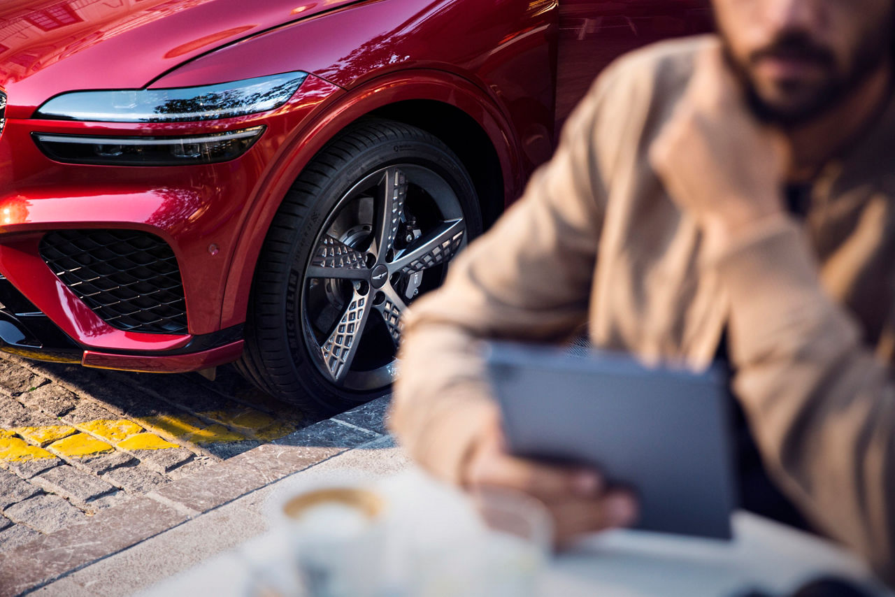 Tyres of a red Genesis GV70 Sport, in the foreground a person with a tablet and coffee