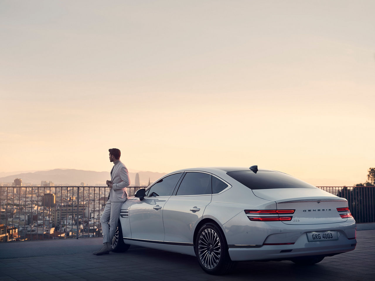Man leaning against a white Genesis G80 and looking at a city in the background