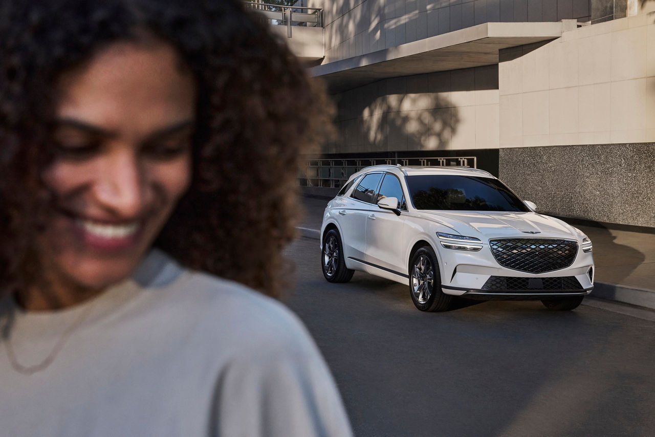 A white Genesis eGV70 parks in front of a house, a smiling woman in the foreground