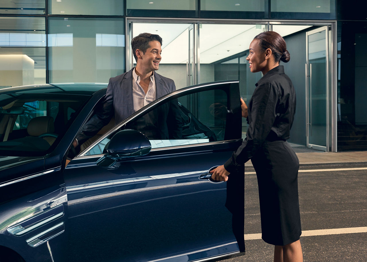 A woman opens the door of a blue Genesis G80 for a man in front of a building