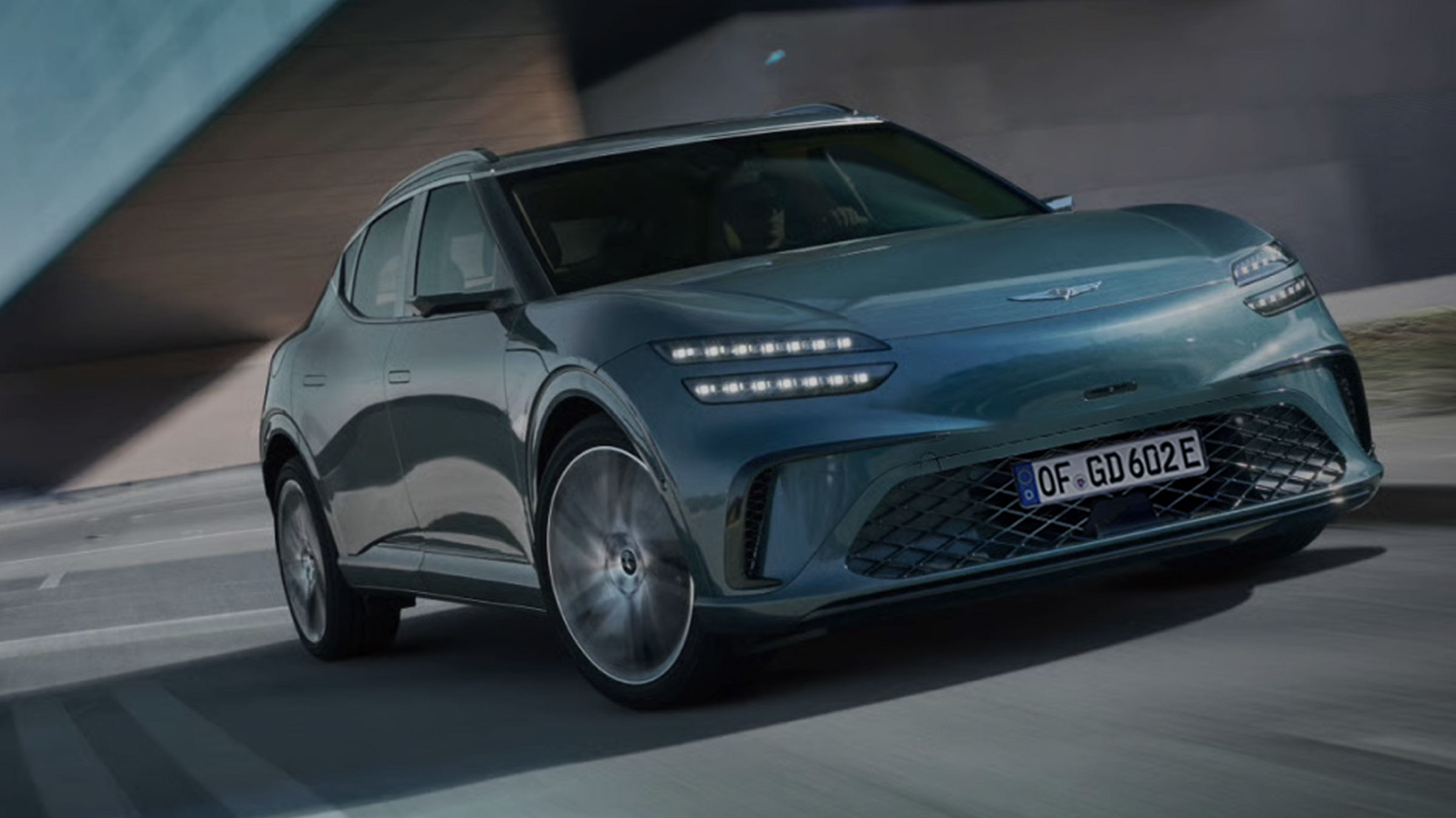  With a modern building in the background, the front view of the GV60 vehicle is seen driving quickly down the highway.