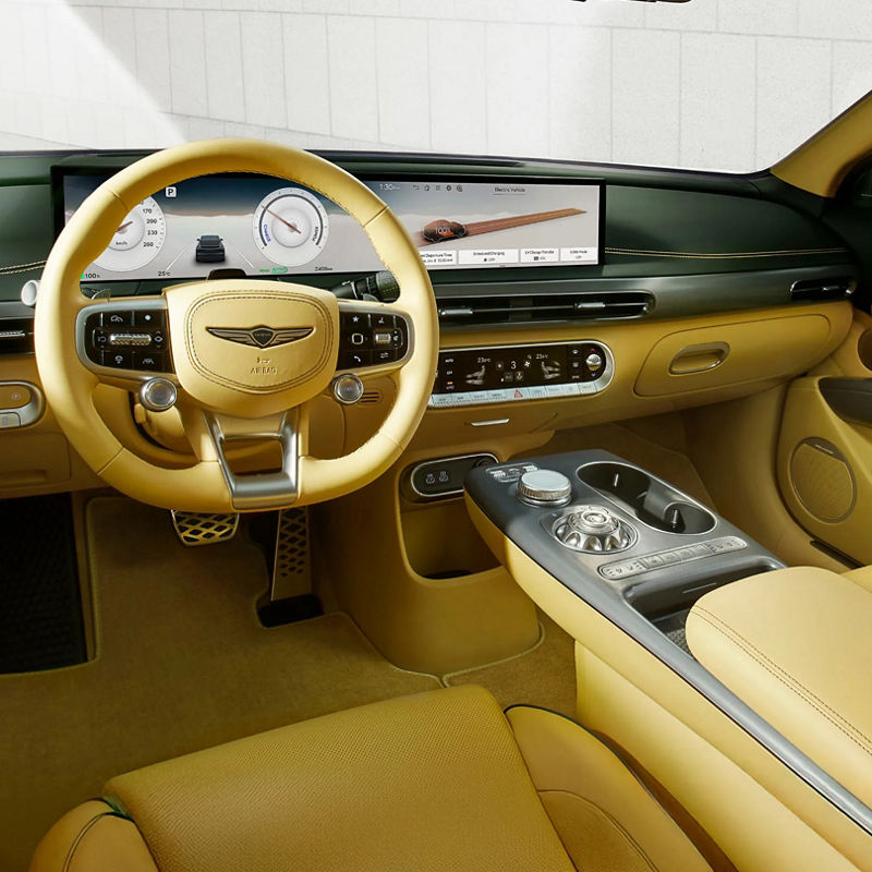  The interior of the front seat of the GV60 vehicle is visible, ranging from various functional buttons located near the vehicle door handle to the steering wheel or display and numerous control buttons.