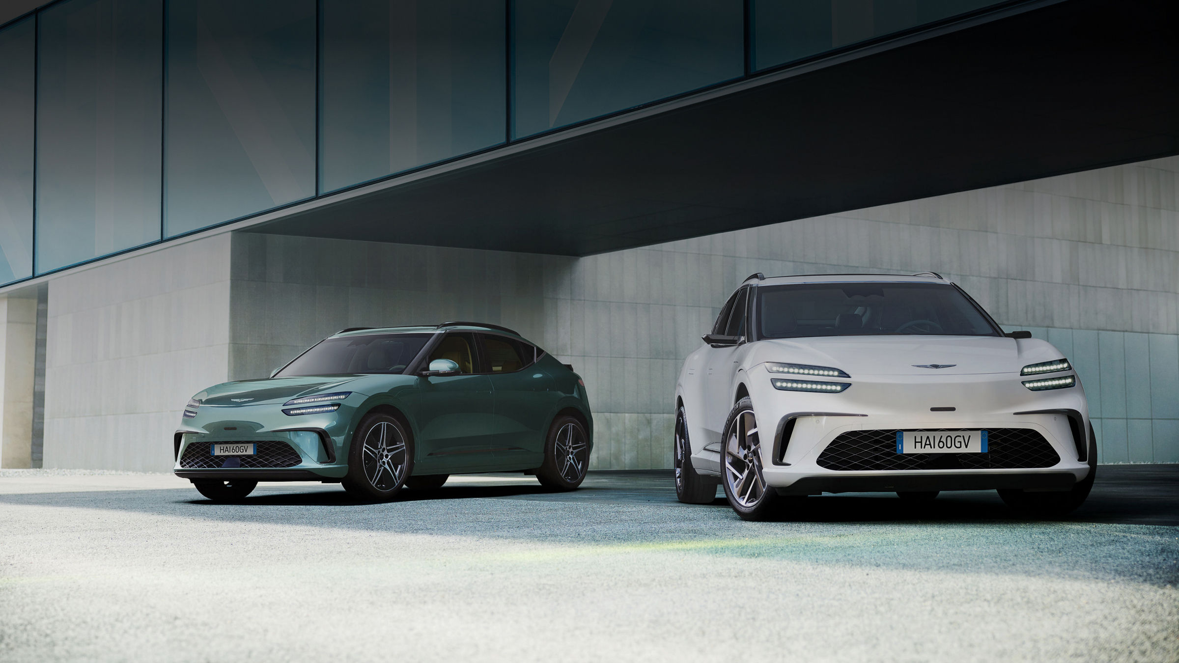 Con un edificio moderno sullo sfondo, a sinistra c’è una GV60 verde Tromso e a destra una GV60 bianca Uyuni.