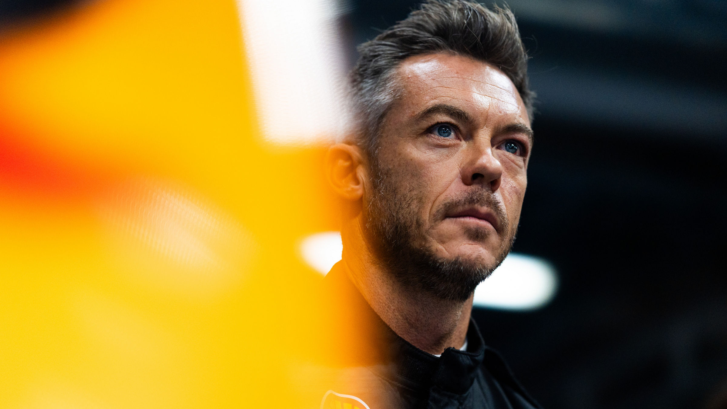 Close-up portrait of André Lotterer, partially framed by an out-of-focus orange race car bodywork in the foreground.