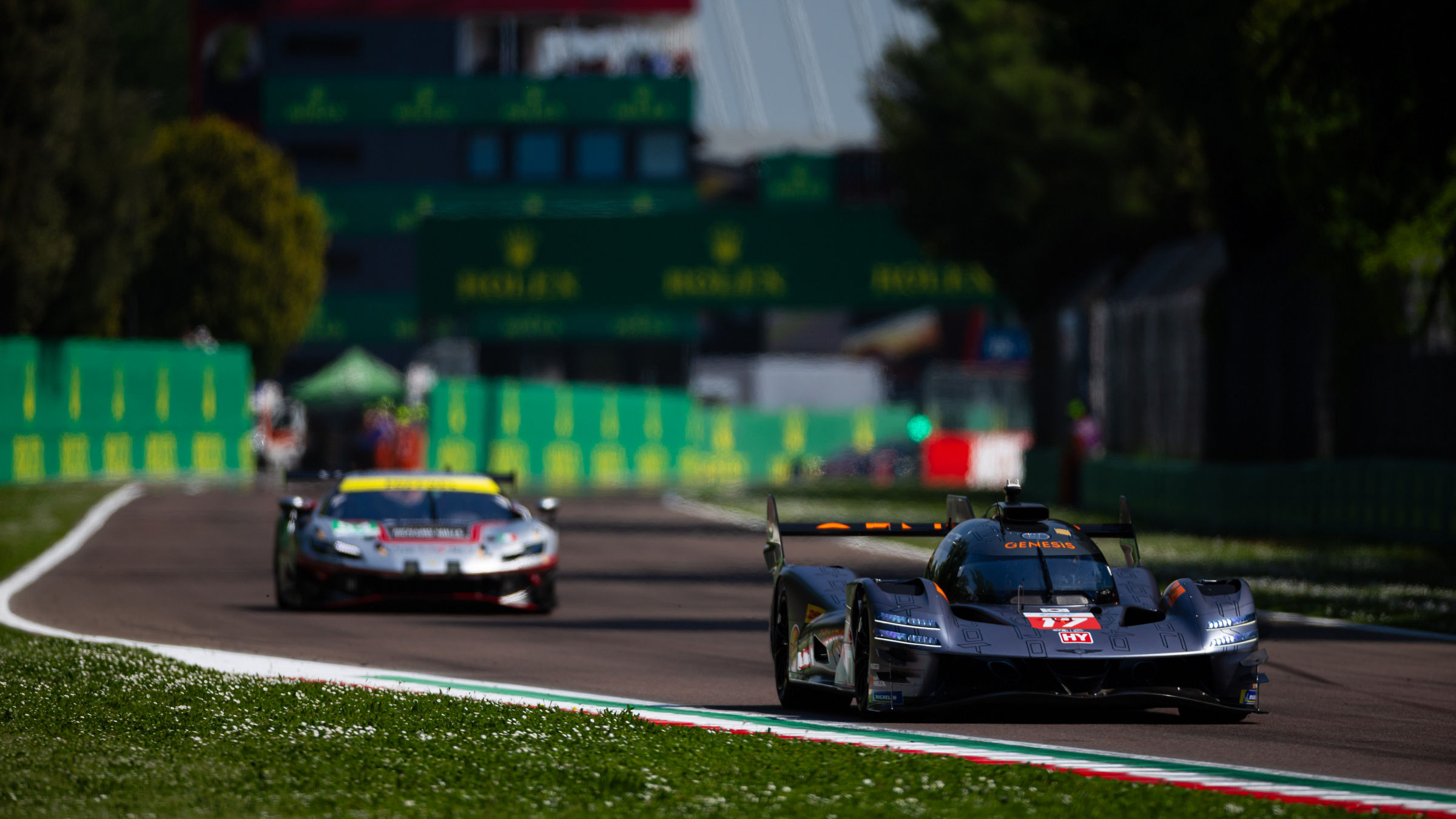 Genesis GRM-001 leading a GT car on a sunlit track with green barriers.