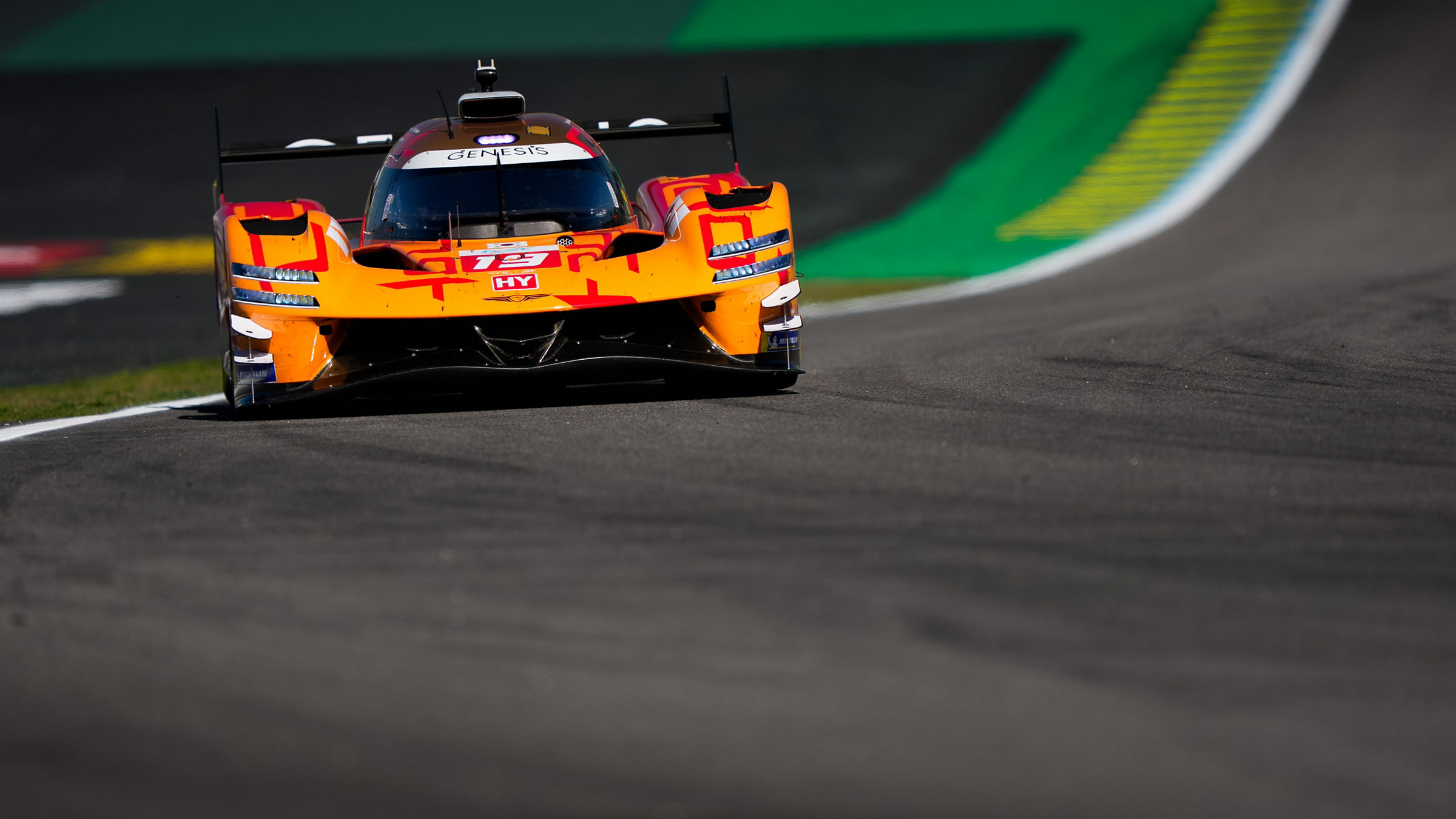 Front-on view of the Genesis GRM-001 cresting a section of the Interlagos circuit, highlighting the prototype’s aerodynamic design and distinctive orange Genesis Magma Racing livery.