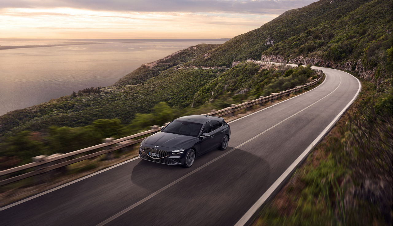 A Genesis G70 drives on a road in the mountains with the sea in the background