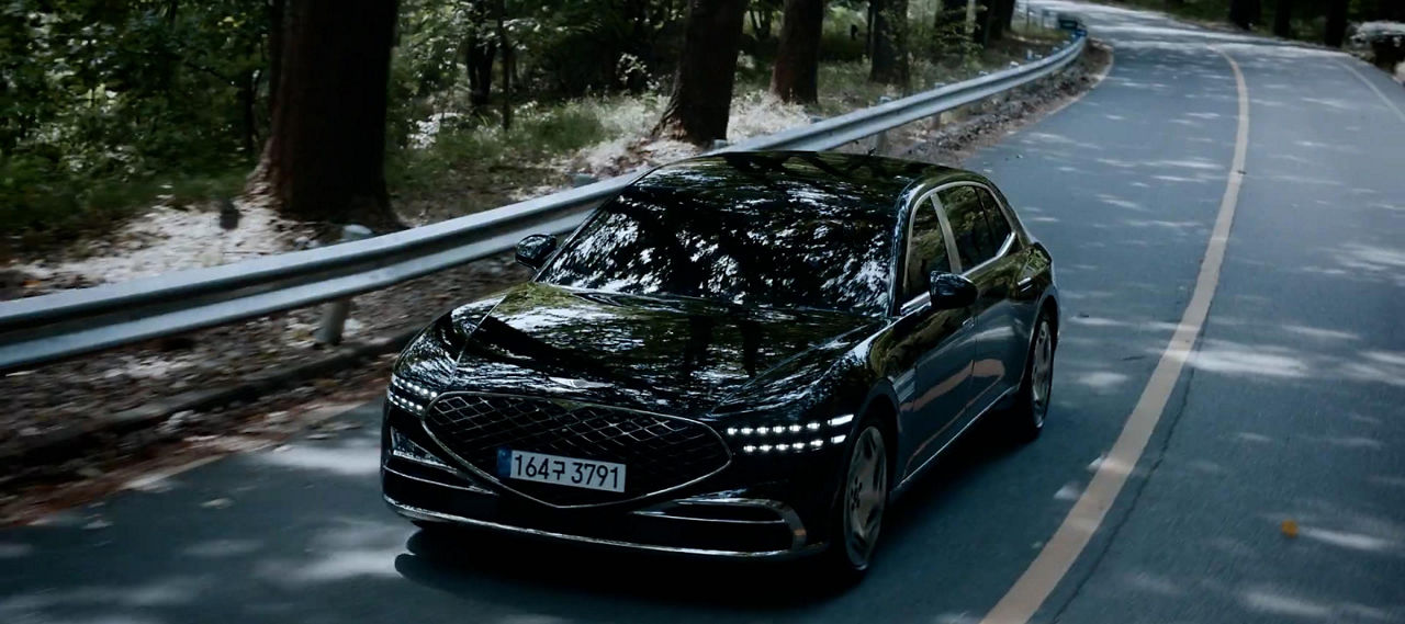 Black Genesis G90 from the front driving on a shady road with trees next to it