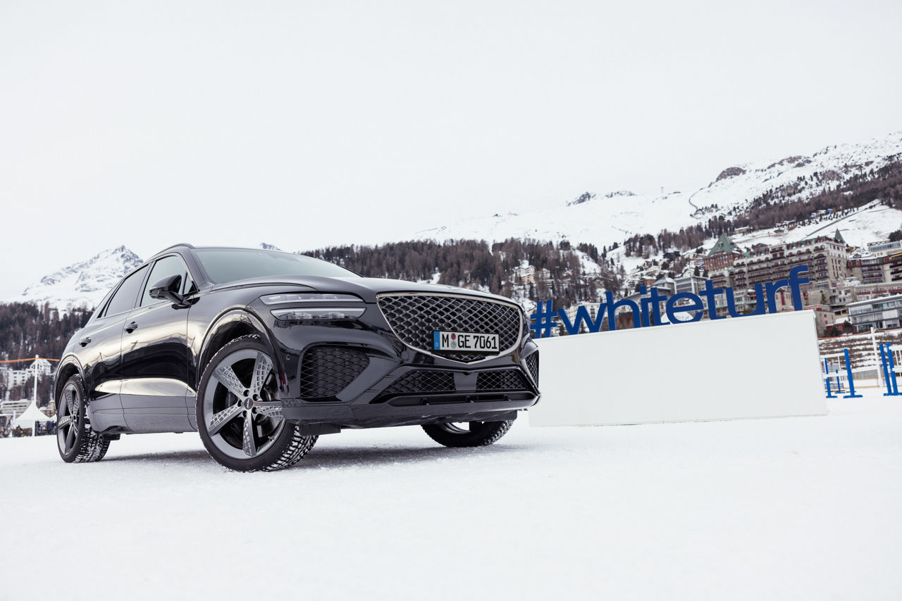 Genesis GV70 noire sur une route enneigée avec des montagnes et des maisons en arrière-plan