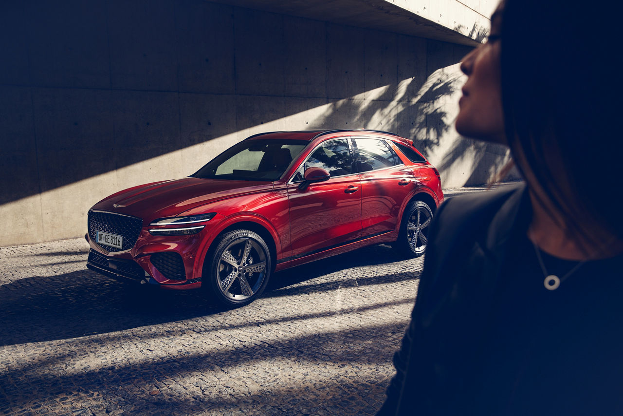 A woman in the foreground looks over her shoulder at a red Genesis GV70