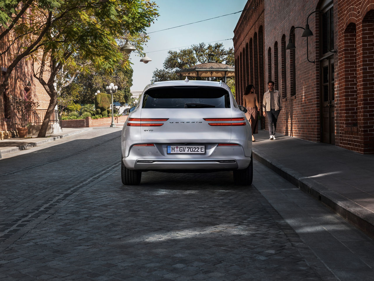 Silver Genesis GV70 from behind in the city next to 2 people on a footpath