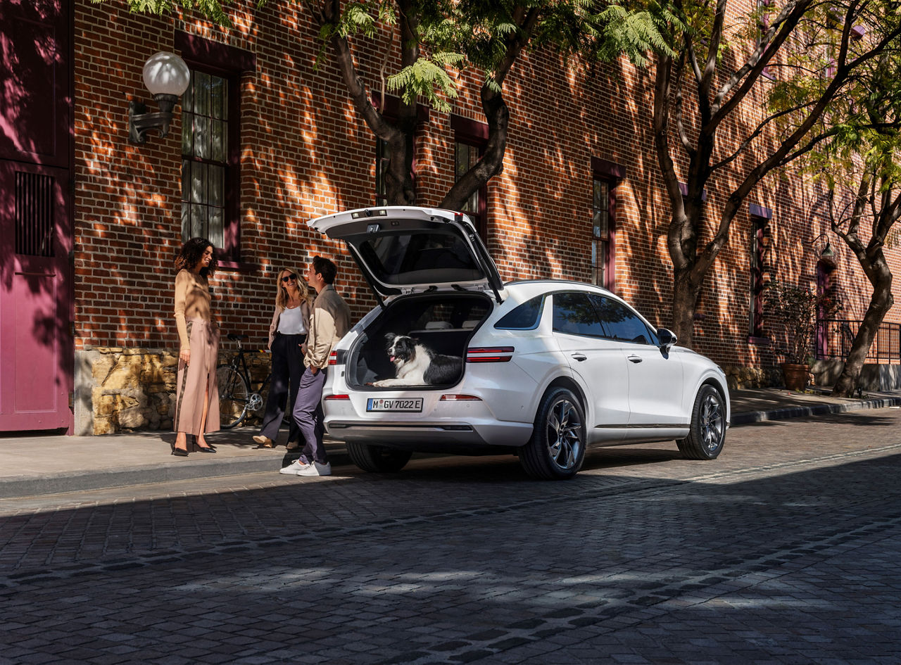 Trois personnes se tiennent à côté d'une Genesis Electrified GV70 blanche avec le coffre ouvert et un chien à l'intérieur