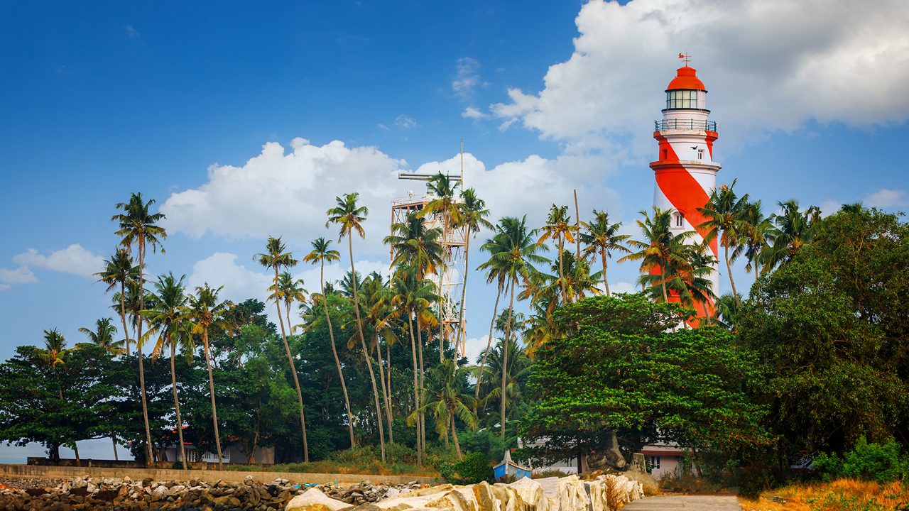 Thangassery red and white stripe Lighthouse on the cliff surrounded by palm trees and big sea waves on the Kollam beach. Kerala, India