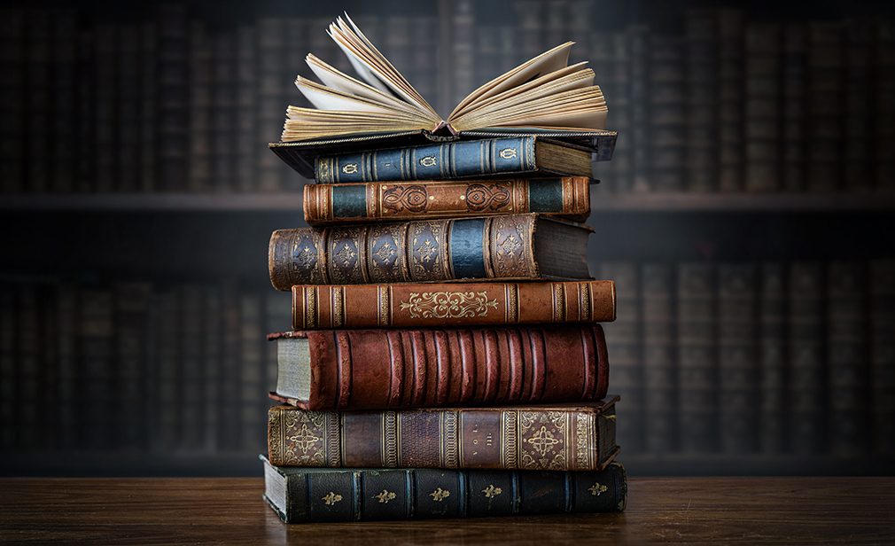 A stack of old books on table against background of bookshelf in library. Ancient books as a symbol of knowledge, history, memory and information. Conceptual background on education, literature topics