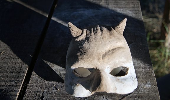 Horned mask. Mask of the devil. A photo of a white mask made of papier-mache. The mask lies on an old wooden table. Sun rays, side lighting, summer day. Handmade in the process.