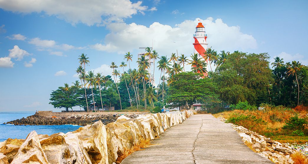 Thangassery red and white stripe Lighthouse on the cliff Quay breakwater surrounded by palm trees and big sea waves on the Kollam beach. Kerala, India