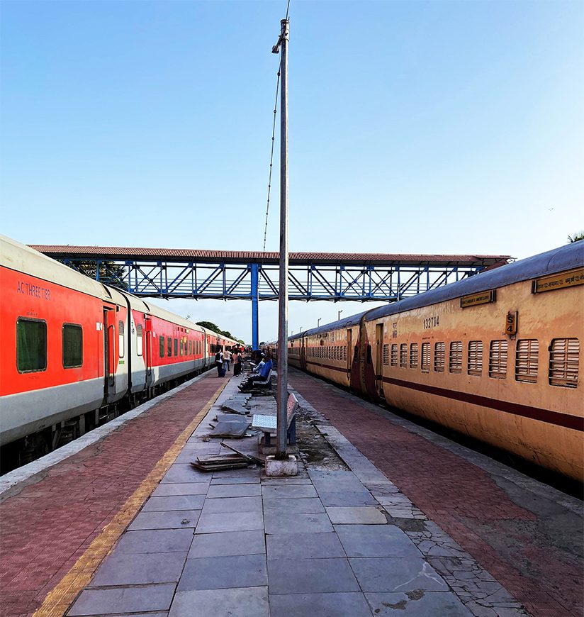 At a rural railway station in Gujarat, India, two Indian railway trains stand facing each other with a diverse tapestry of passengers. People using bridge to arrive and exit the railway station.