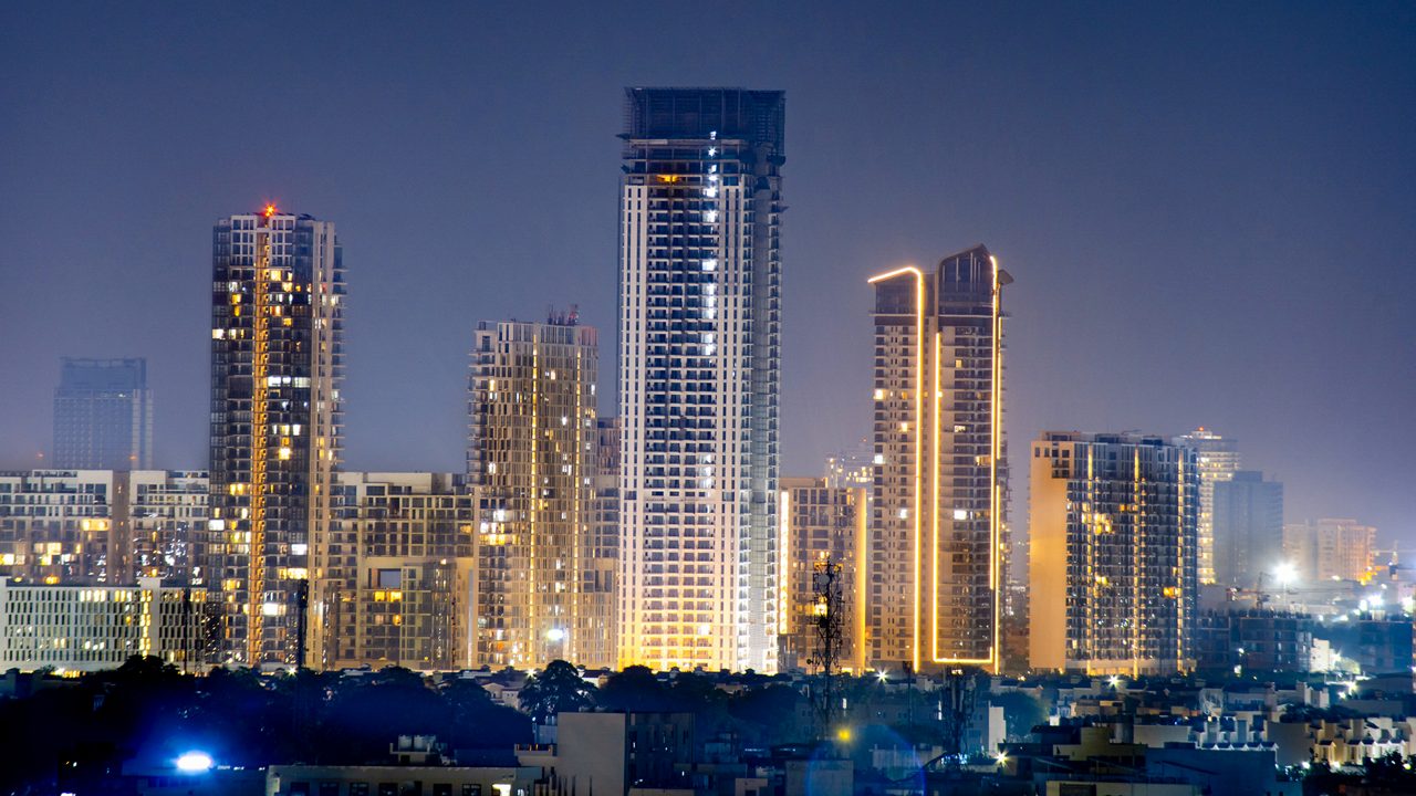 High rise multi story skyscrapers lit up at night with small houses in the foreground at night in gurgaon delhi. Shows the rapid pace of development of the real estate sector with property, offices