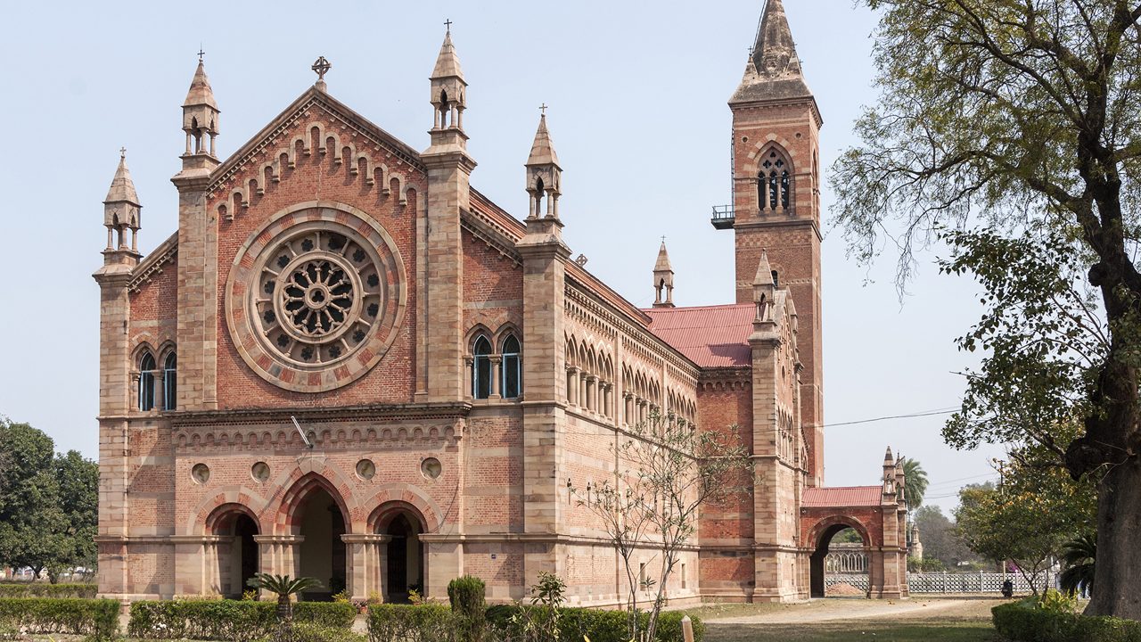 India Kanpur: All Souls Church in its park with tower and rose-window. Classic dominant red brick cathedral building set in green environment and under blue skies.