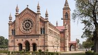 India Kanpur: All Souls Church in its park with tower and rose-window. Classic dominant red brick cathedral building set in green environment and under blue skies.