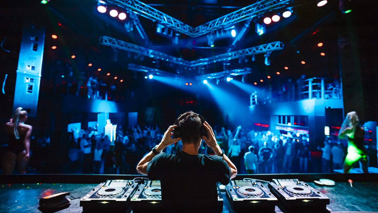 DJ with headphones at night club party under the blue light and people crowd in background