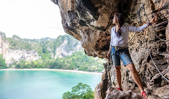 Confidence Asian woman climber with safety rope climbing high up on rocky mountain at tropical island in summer. Healthy and strong female enjoy active lifestyle and extreme sport in holiday vacation.