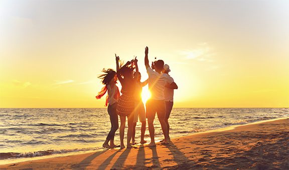 group of happy young people dancing at the beach on beautiful summer sunset