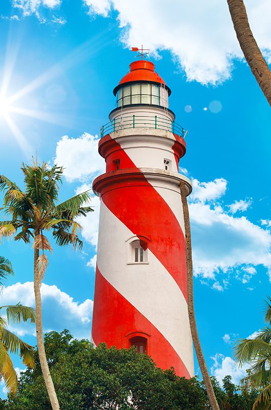 Thangassery red and white stripe Lighthouse on the cliff surrounded by palm trees and big sea waves sun rays on the Kollam beach. Kerala, India