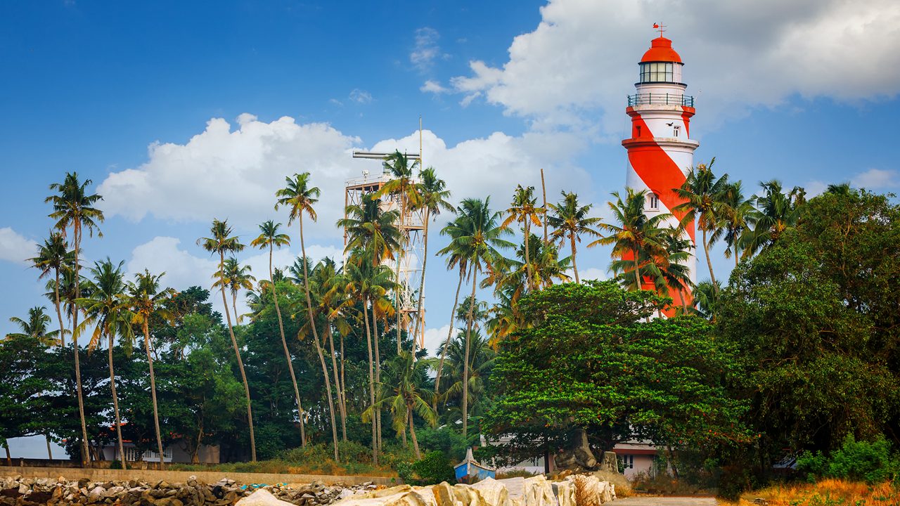 Thangassery red and white stripe Lighthouse on the cliff surrounded by palm trees and big sea waves on the Kollam beach. Kerala, India