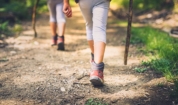 Children hiking in mountains or forest with sport hiking shoes. Girls or boys are walking trough forest path wearing mountain boots and walking sticks. Frog perspective with focus on the shoes.