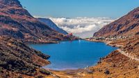 A blue water Tsomgo lake surrounded by desert brown mountains is located high above clouds. The winding road to the distant village leads a way to heaven in cumulus clouds and blue sky. Sikkim, India