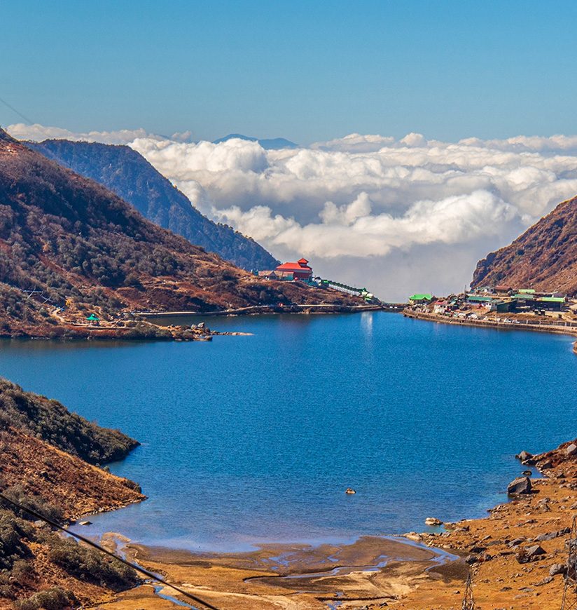 A blue water Tsomgo lake surrounded by desert brown mountains is located high above clouds. The winding road to the distant village leads a way to heaven in cumulus clouds and blue sky. Sikkim, India