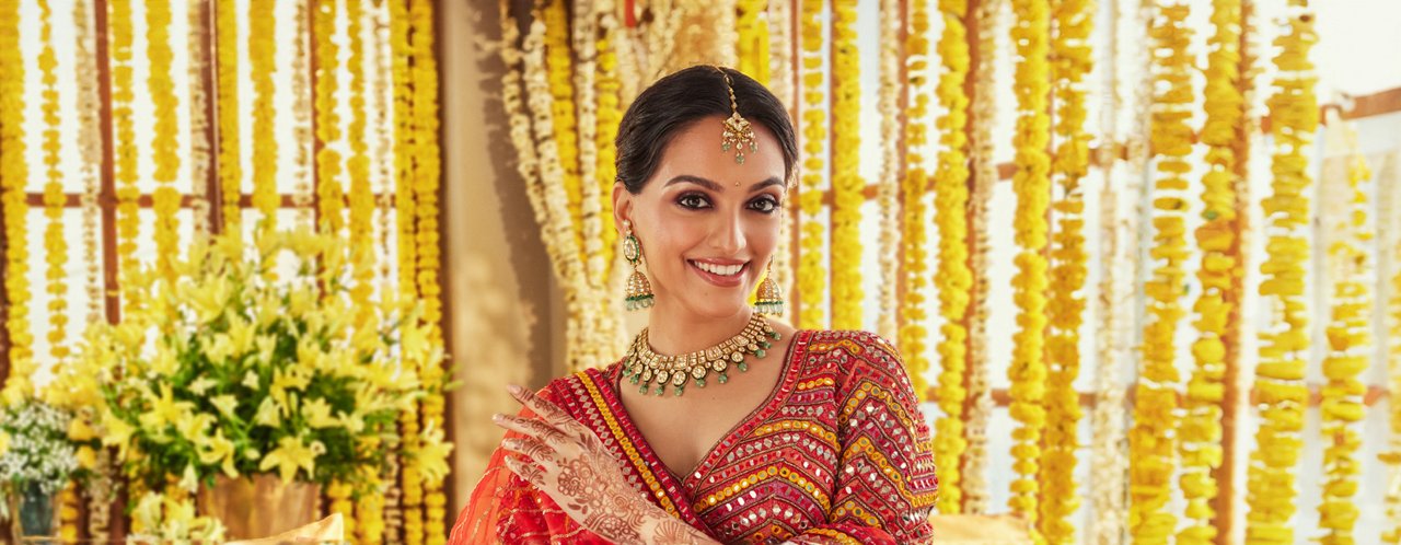 Radiant bride in red saree with elaborate gold jewellery, smiling amidst floral backdrop