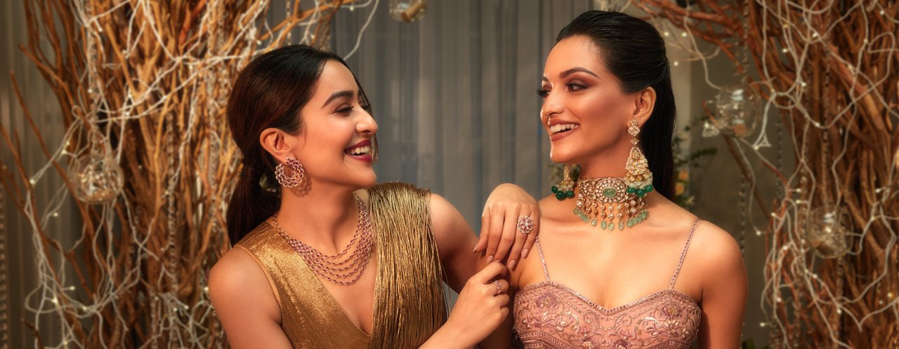 Two women laughing, one in gold attire with circular earrings, the other in pink with green jeweled necklace