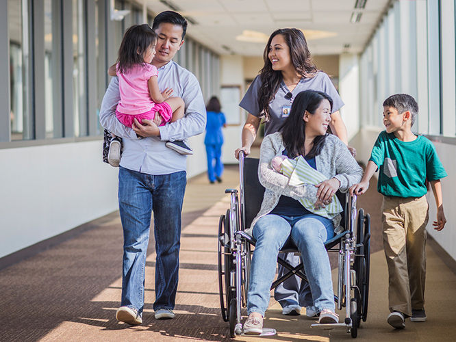 A family is seen walking through a brightly lit hospital corridor. A woman in a wheelchair holds a baby, accompanied by a man carrying a young child and a boy walking beside them. The setting suggests a supportive and caring environment, with medical staff visible in the background.